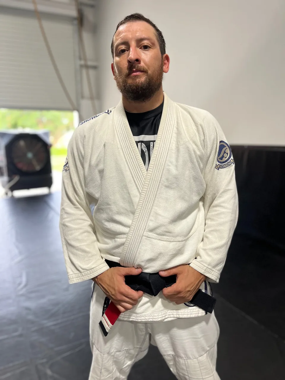 Justin Wood in a white gi with his black belt, standing on the mat at the academy.