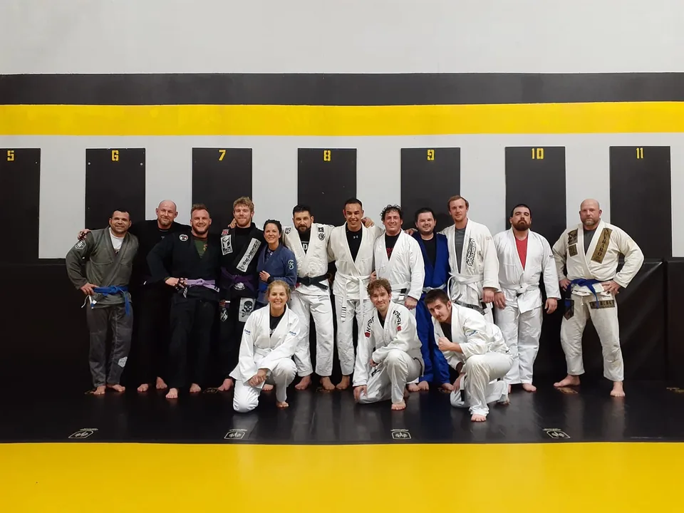 A training group in gis and rash guards posed together on the black and yellow mat in front of numbered wall panels, Felipe Soares standing at the far left in a grey gi holding a blue belt.