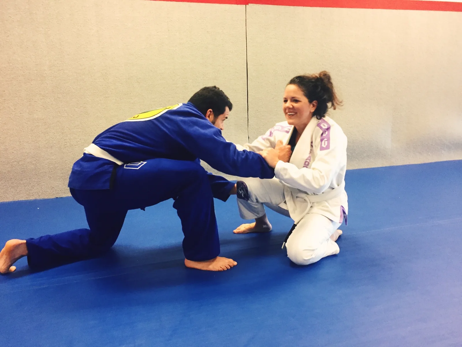 Felipe Soares in a blue gi drilling a grip and knee-slide entry with his wife Jeralin Soares, who kneels in a white gi with a white belt, both smiling on the blue mat.