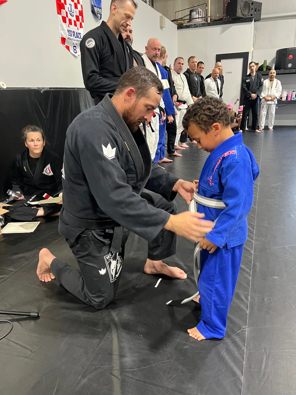 Justin Wood kneeling in a black gi to tie a new white belt on a young student in a blue gi while the class watches.