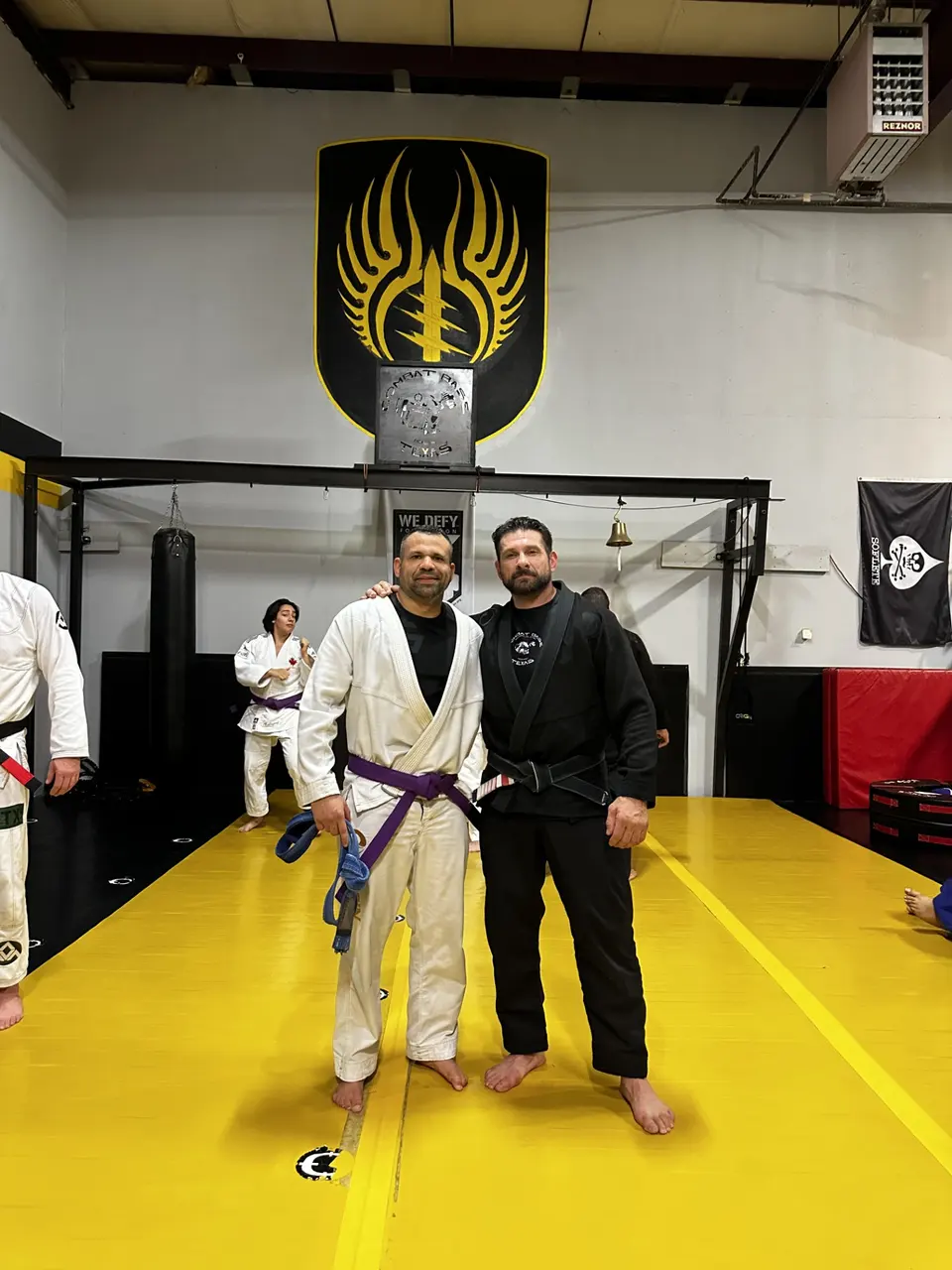Felipe Soares, organizer of Train My Hands for War, wearing a white gi with his purple belt tied at the waist, standing on the mat beside his coach Allan Shebaro in a black gi.