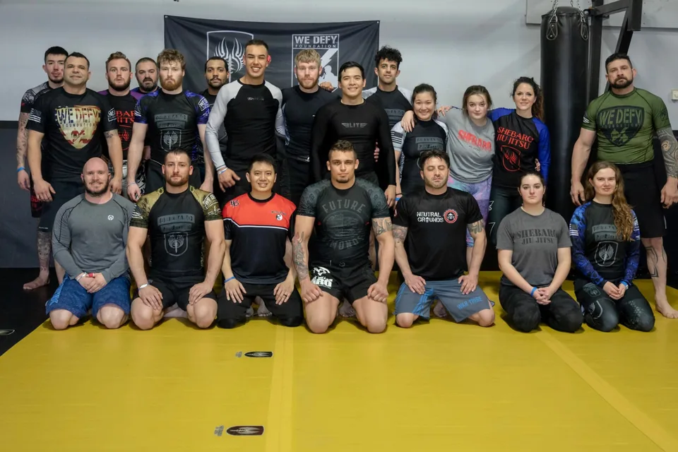 A large no-gi class in rash guards standing and kneeling on the yellow mat in front of a We Defy Foundation banner at Shebaro Jiu Jitsu.