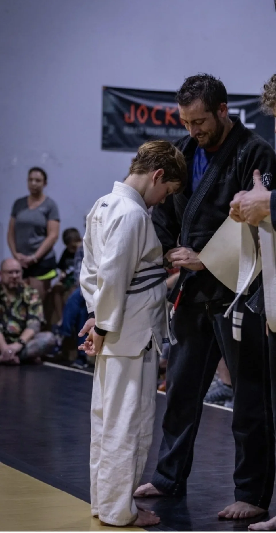 Justin Wood in a black gi tying a new belt on a youth student in a white gi while families watch from the edge of the mat.
