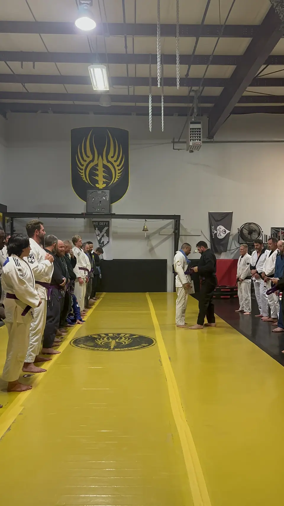 Felipe Soares standing in a white gi on the yellow mat at Shebaro BJJ while Allan Shebaro presents him with his new purple belt, teammates lined up along both sides of the mat.