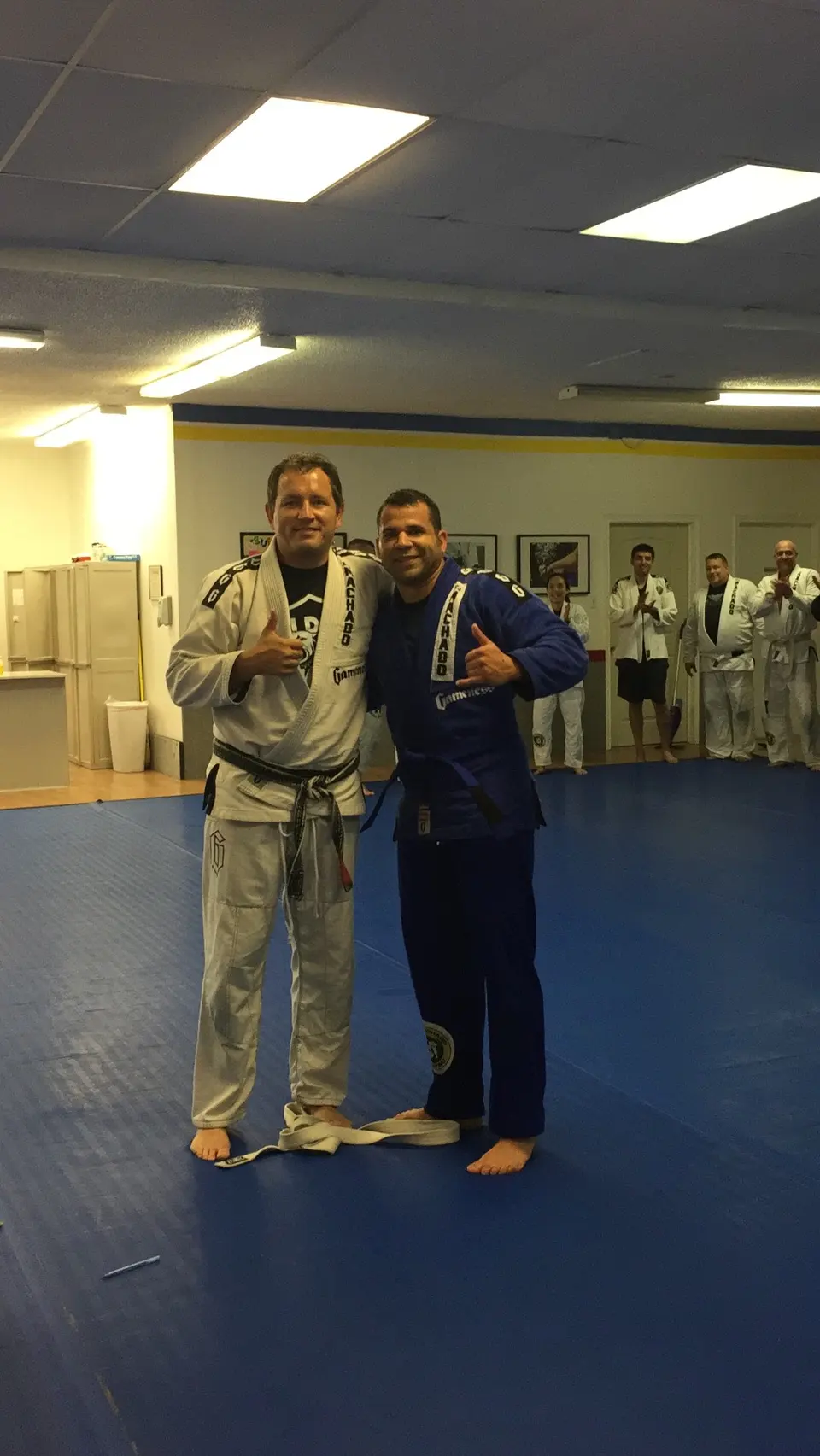 Felipe Soares in a blue gi wearing his new blue belt, standing beside coach Michael Medley in a white gi and black belt on the mat at RCJ Machado in The Colony, Texas, with students applauding in the background.