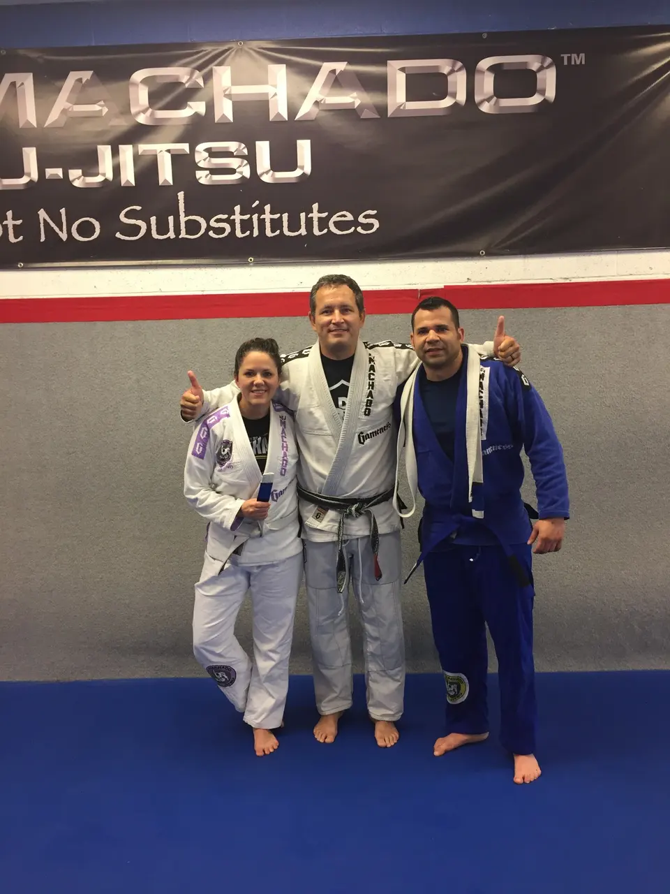 Felipe Soares in a blue gi with his blue belt and Jeralin Soares in a white and purple gi, standing on either side of coach Michael Medley in a white gi and black belt under the RCJ Machado Jiu-Jitsu banner.