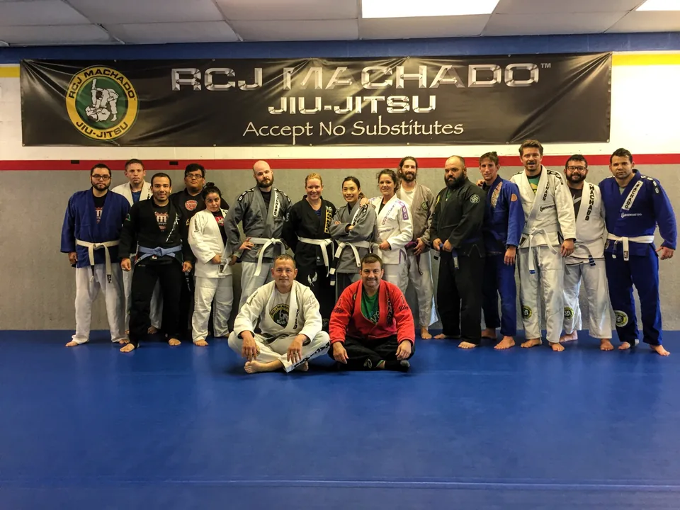 Students and coaches in gis posed on the blue mat beneath a large RCJ Machado Jiu-Jitsu banner reading Accept No Substitutes.