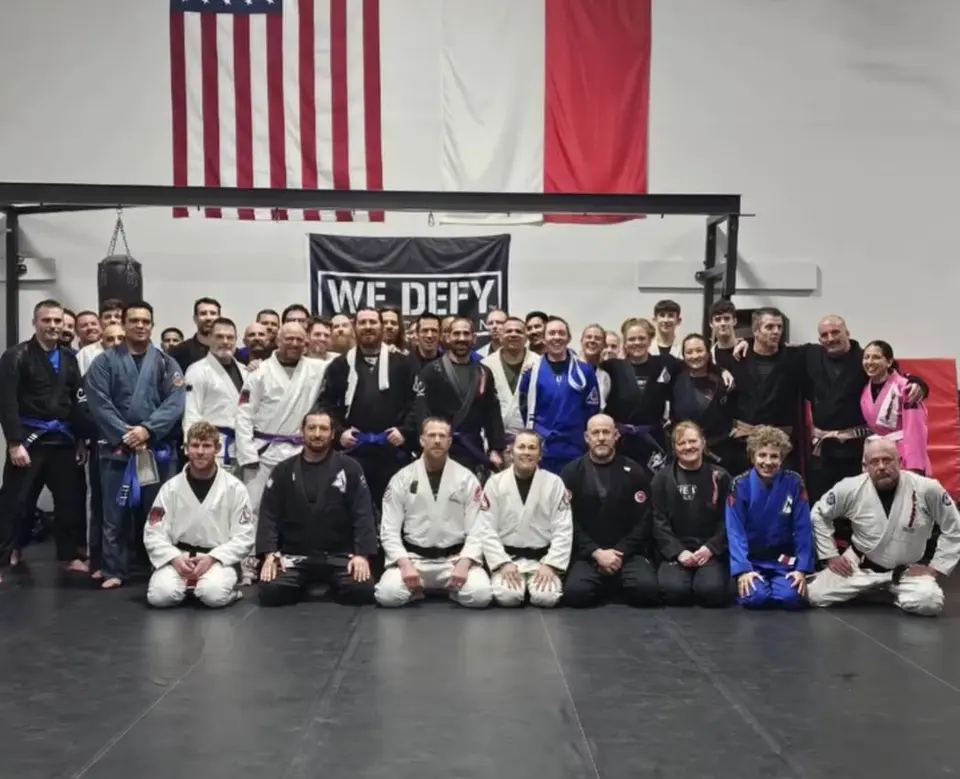 A large group of students and coaches in white, blue, and black gis posed together on the mat at 3 Embers Grappling Academy in McKinney, Texas, with an American flag on the wall behind them.