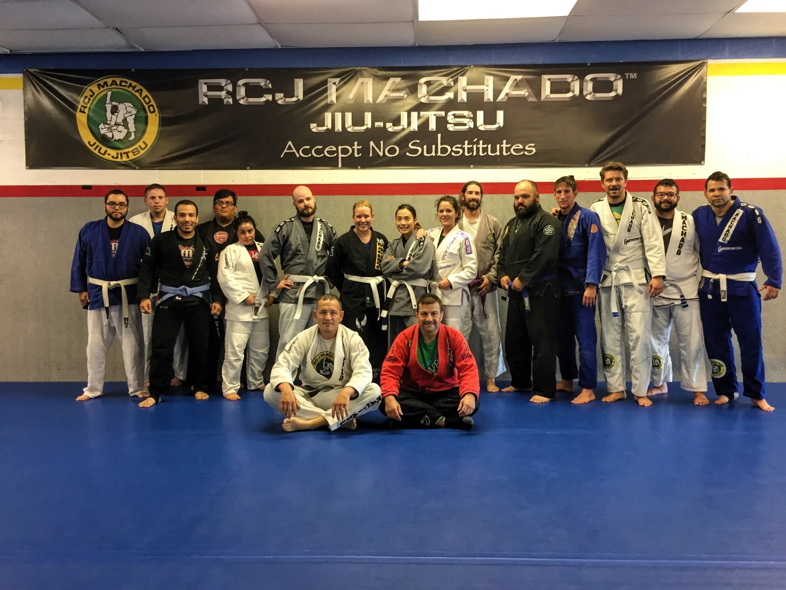 Students and coaches in gis posed on the blue mat beneath a large RCJ Machado Jiu-Jitsu banner reading Accept No Substitutes.