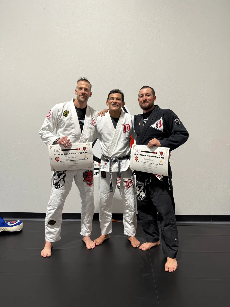 Justin Wood in a black gi holding an IBJJF second degree black belt certificate beside Professor Alex Martins and Mike Anderson.