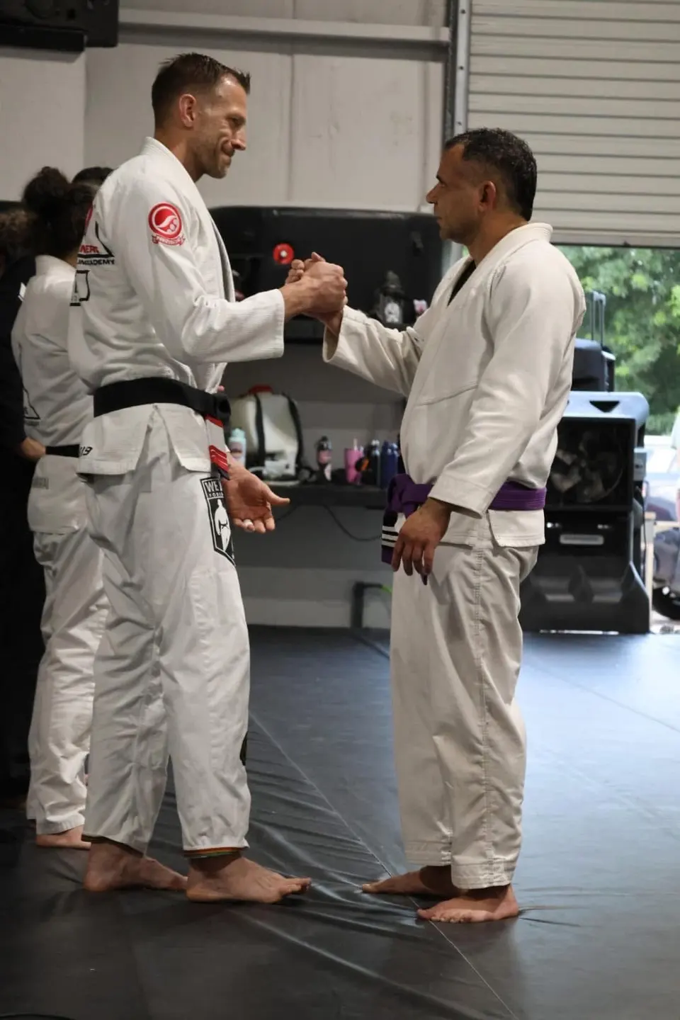 Felipe Soares in a white gi with a purple belt shaking hands with coach Michael Anderson in a white gi and black belt after receiving his stripe at 3 Embers Grappling Academy.