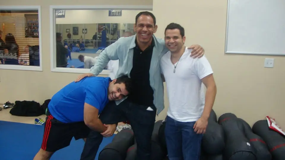 Felipe Soares in a white shirt laughing alongside teammates and coaches beside the mats at Minotauro BJJ during a training session.