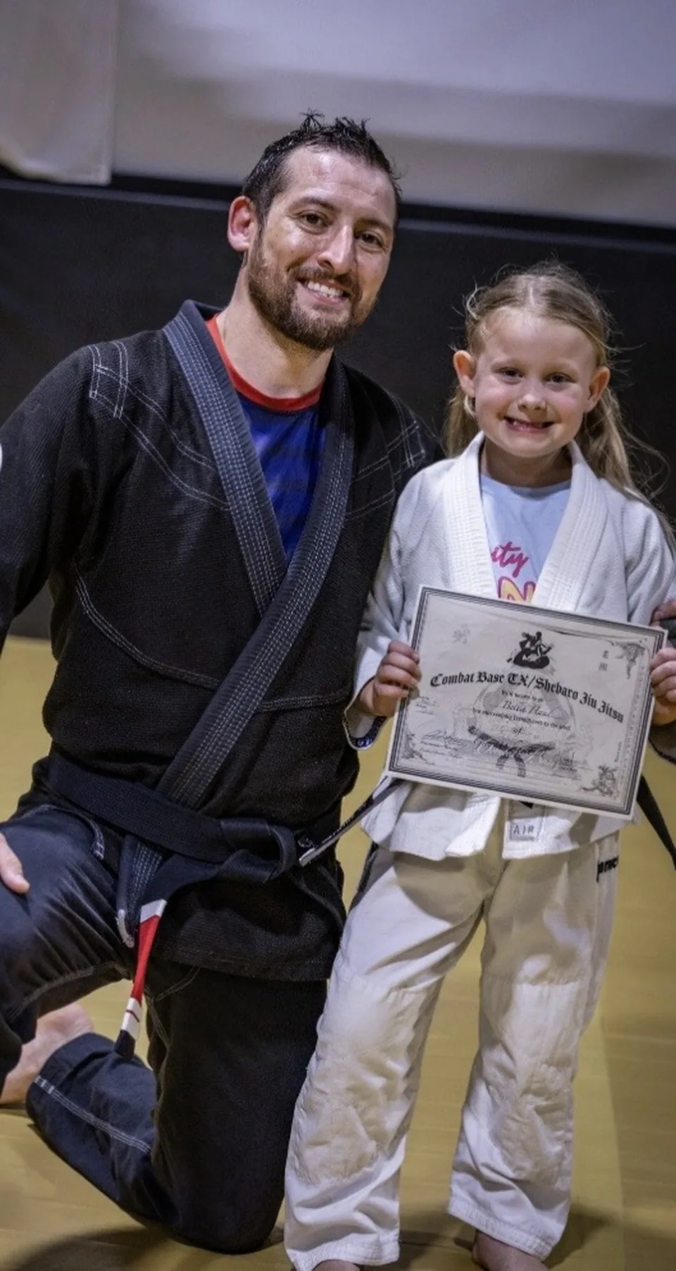 Justin Wood kneeling in a black gi beside a young student in a white gi who is holding a Combat Base TX / Shebaro Jiu Jitsu certificate.
