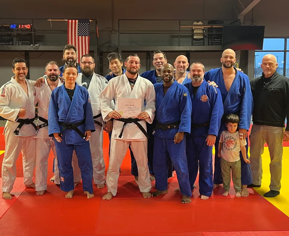 Justin Wood in a white judo gi holding his Shodan certificate, surrounded by teammates and coaches on the mat.