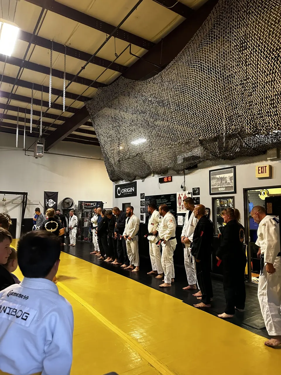 Students and coaches lined up in gis along the edge of the yellow mat at Shebaro BJJ for the start of class beneath a camouflage net.