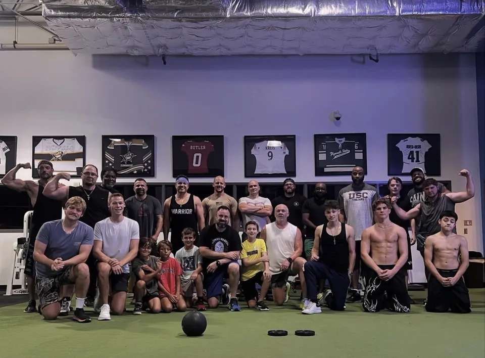 About a dozen men and teenage boys in workout clothes standing shoulder to shoulder, smiling, on the gym floor after a Lifted Faith 5 AM Club Saturday workout in Frisco, Texas