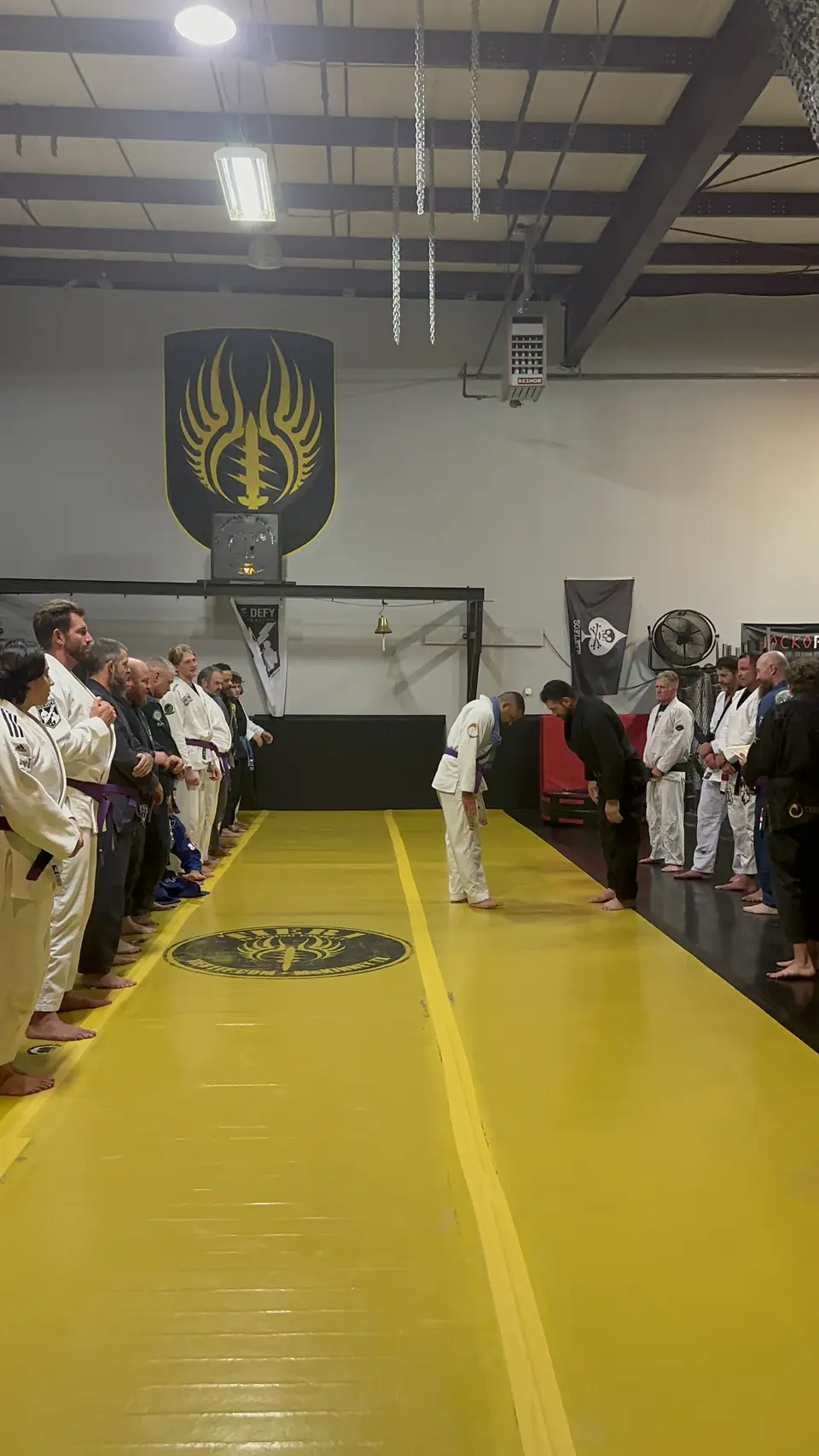 Felipe Soares in a white gi with his newly tied purple belt bowing to coach Allan Shebaro in a black gi on the yellow mat at Shebaro BJJ.