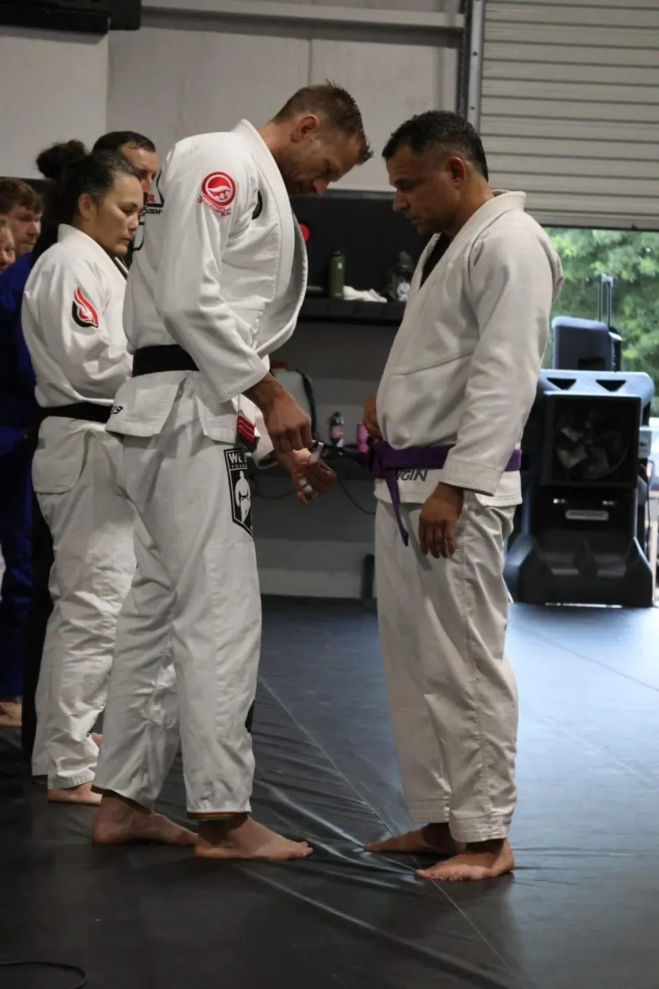 Coach Michael Anderson in a white gi and black belt adding a stripe to Felipe Soares' purple belt while Felipe stands at attention in a white gi at 3 Embers Grappling Academy.