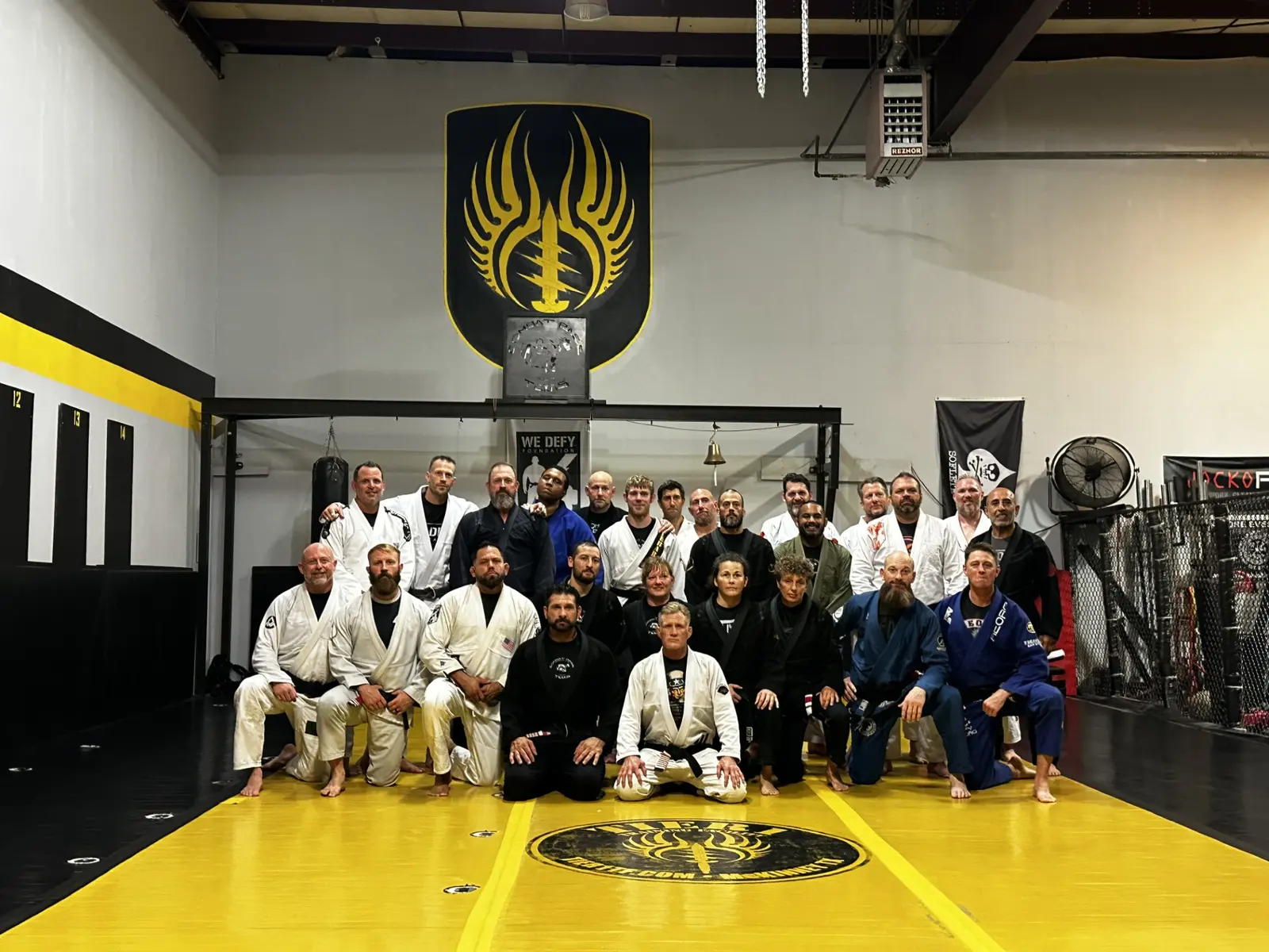 A full class of students and coaches in white, black, and blue gis posed together on the yellow mat under the Texas Combat Base crest at Shebaro BJJ.