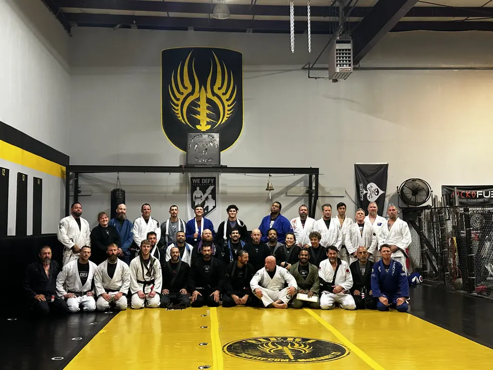 A large group photo of Shebaro BJJ students and coaches kneeling and standing on the yellow mat beneath the gold Texas Combat Base crest.