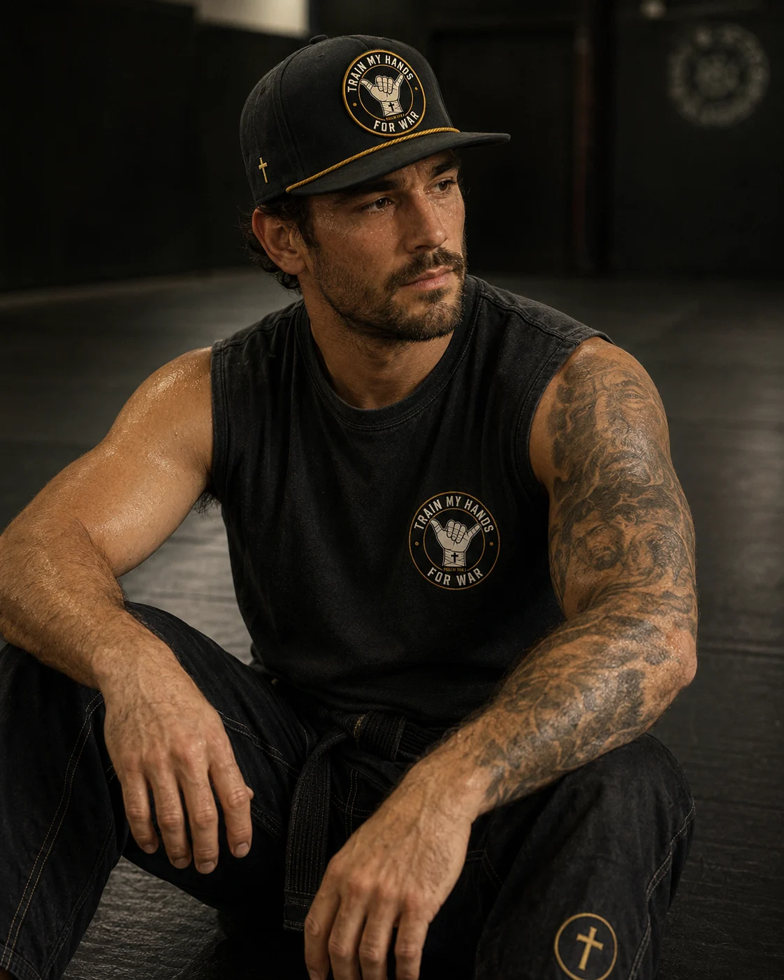 Member in a washed-black sleeveless TMHFW shirt and rope hat, towel over his shoulder, mat-side