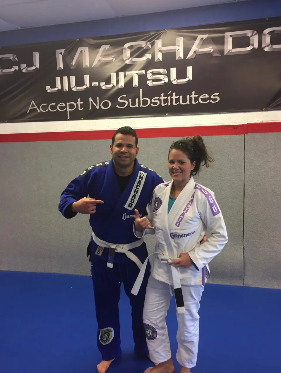 Felipe Soares in a blue gi with a white belt pointing at his RCJ Machado gi patch, next to Jeralin Soares in a white and purple gi with a white belt, under the RCJ Machado Jiu-Jitsu banner.